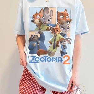 K&ouml;nnte beinhalten: Hellblaues T-Shirt mit einem Comic-Strip-Design mit Charakteren aus "Zootopia 2". Die Grafik zeigt Judy Hopps, Nick Wilde und andere Charaktere. Der Text "ZOOTOPIA 2" ist darunter gedruckt.