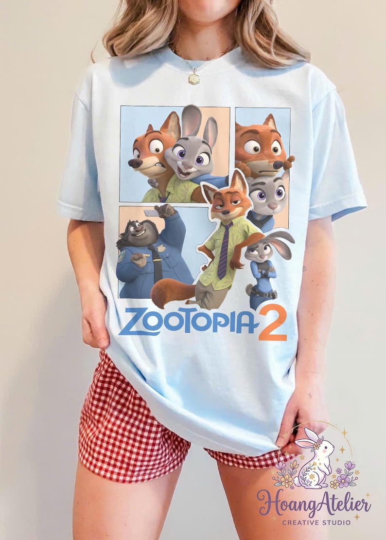 K&ouml;nnte beinhalten: Hellblaues T-Shirt mit einem Comic-Strip-Design mit Charakteren aus "Zootopia 2". Die Grafik zeigt Judy Hopps, Nick Wilde und andere Charaktere. Der Text "ZOOTOPIA 2" ist darunter gedruckt.