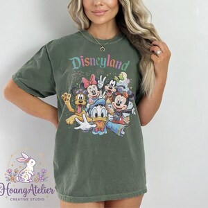 May include: Olive green t-shirt featuring the word "Disneyland" in colorful text above a graphic of Disney characters including Mickey Mouse, Minnie Mouse, Donald Duck, and Goofy. The shirt has short sleeves.