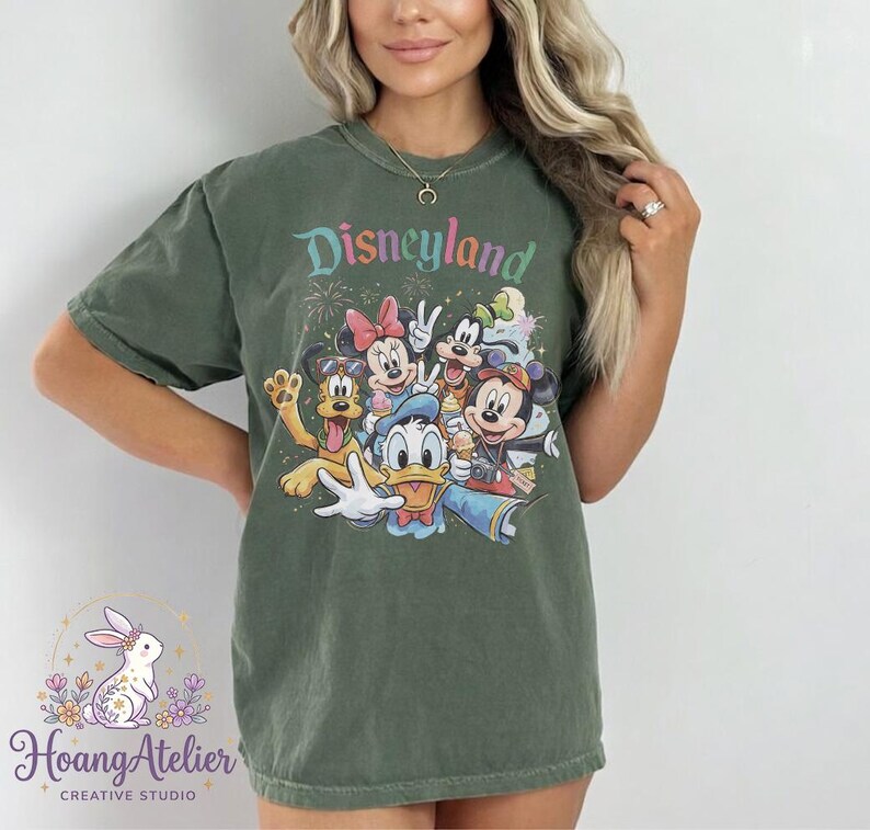 May include: Olive green t-shirt featuring the word "Disneyland" in colorful text above a graphic of Disney characters including Mickey Mouse, Minnie Mouse, Donald Duck, and Goofy. The shirt has short sleeves.