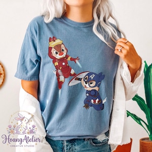 May include: Blue t-shirt featuring cartoon characters dressed as superheroes. One character is in a red Iron Man suit, and the other is in a Captain America suit. The shirt has the Hoang Atelier logo at the bottom.