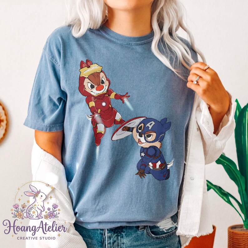 May include: Blue t-shirt featuring cartoon characters dressed as superheroes. One character is in a red Iron Man suit, and the other is in a Captain America suit. The shirt has the Hoang Atelier logo at the bottom.