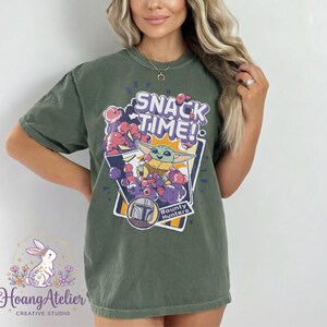 Puede incluir: Camiseta verde oliva con un gr&aacute;fico de Grogu de The Mandalorian y el texto "SNACK TIME!". El dise&ntilde;o incluye un casco de cazarrecompensas y detalles morados y rojos. La camiseta es de estilo casual.