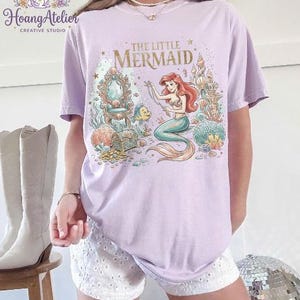 May include: A light purple t-shirt featuring a colorful illustration of Ariel from "The Little Mermaid." The design includes Ariel, Flounder, a treasure chest, and a castle. The text "THE LITTLE MERMAID" is printed in gold.