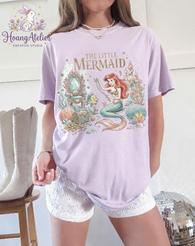 May include: A light purple t-shirt featuring a colorful illustration of Ariel from "The Little Mermaid." The design includes Ariel, Flounder, a treasure chest, and a castle. The text "THE LITTLE MERMAID" is printed in gold.