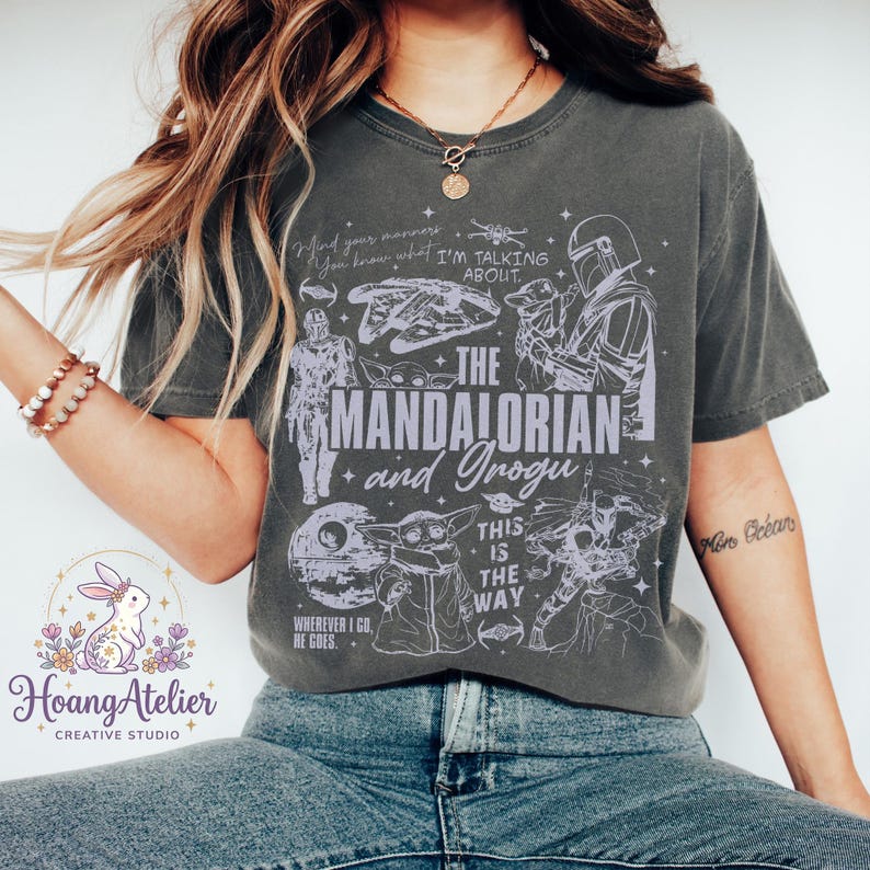 Puede incluir: Camiseta gris oscuro con un dise&ntilde;o gr&aacute;fico con personajes y texto de la serie "The Mandalorian". El dise&ntilde;o incluye el texto "The Mandalorian and Grogu", "This is the Way" y otras frases e im&aacute;genes. La camiseta est&aacute; siendo usada.