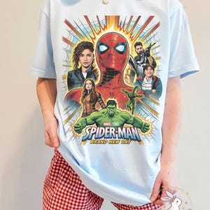 May include: Light blue t-shirt featuring a colorful graphic of Spider-Man and other characters. The design includes the text "SPIDER-MAN BRAND NEW DAY". The shirt is a crew neck and has short sleeves.