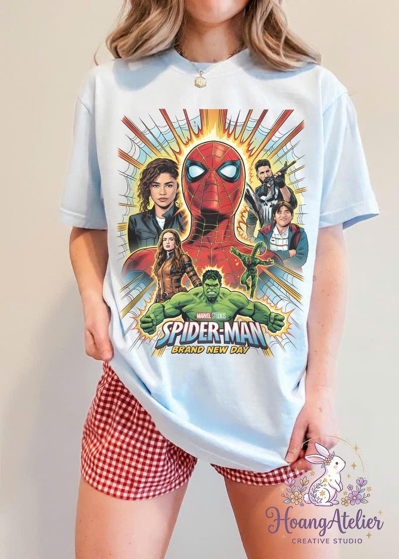 May include: Light blue t-shirt featuring a colorful graphic of Spider-Man and other characters. The design includes the text "SPIDER-MAN BRAND NEW DAY". The shirt is a crew neck and has short sleeves.