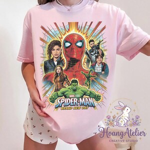 May include: Light pink t-shirt featuring a colorful graphic of Spider-Man and other Marvel characters. The design includes the text "SPIDER-MAN BRAND NEW DAY" and images of Zendaya, Hulk, and others. The shirt is a crew neck.