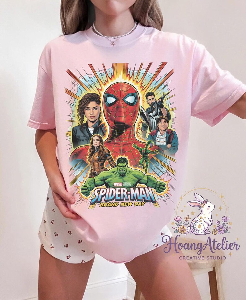 May include: Light pink t-shirt featuring a colorful graphic of Spider-Man and other Marvel characters. The design includes the text "SPIDER-MAN BRAND NEW DAY" and images of Zendaya, Hulk, and others. The shirt is a crew neck.