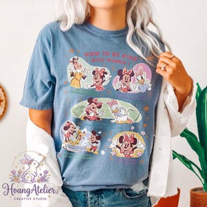 May include: Blue t-shirt with a graphic of Minnie Mouse and friends, and the text "HOW TO BE KIND WITH MINNIE". The design includes illustrations of Minnie Mouse with phrases like "Say hello everyone" and "Helpful makes the world go round".