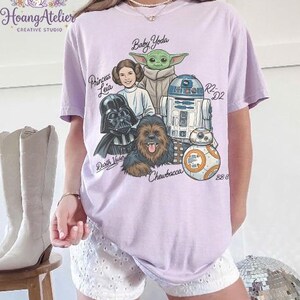 May include: A light purple t-shirt featuring a graphic of Star Wars characters including Baby Yoda, Princess Leia, Darth Vader, Chewbacca, R2-D2, and BB-8. The names of the characters are written above them in a handwritten font. The shirt has short sleeves.