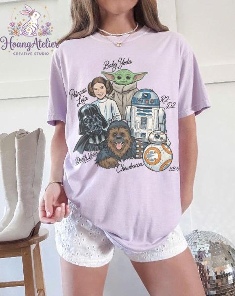 May include: A light purple t-shirt featuring a graphic of Star Wars characters including Baby Yoda, Princess Leia, Darth Vader, Chewbacca, R2-D2, and BB-8. The names of the characters are written above them in a handwritten font. The shirt has short sleeves.