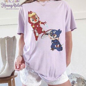 May include: A light purple t-shirt featuring cartoon characters Chip and Dale as Iron Man and Captain America. The shirt has the text "Hoang Atelier CREATIVE STUDIO" and a bunny logo in the upper left corner.
