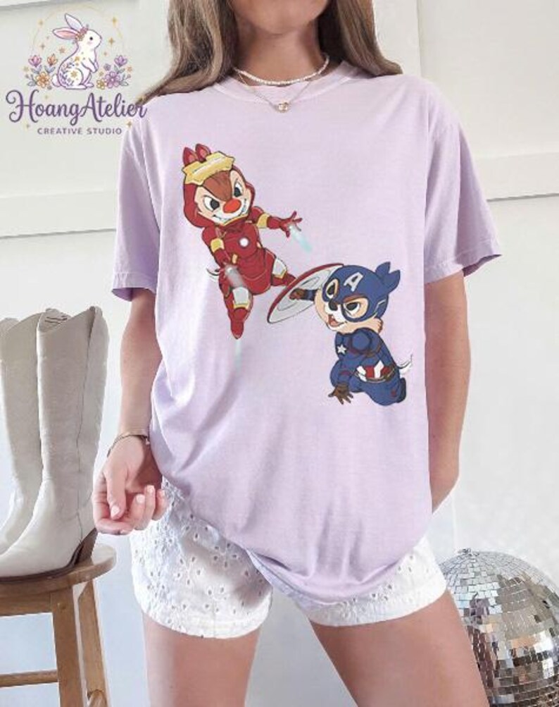 May include: A light purple t-shirt featuring cartoon characters Chip and Dale as Iron Man and Captain America. The shirt has the text "Hoang Atelier CREATIVE STUDIO" and a bunny logo in the upper left corner.