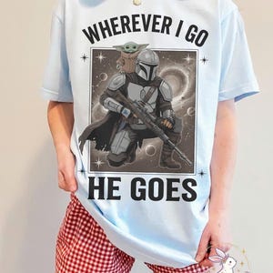 May include: Light blue t-shirt featuring a graphic of The Mandalorian and Grogu, with the text "WHEREVER I GO HE GOES". The design includes a detailed illustration of the characters in a space-themed background.