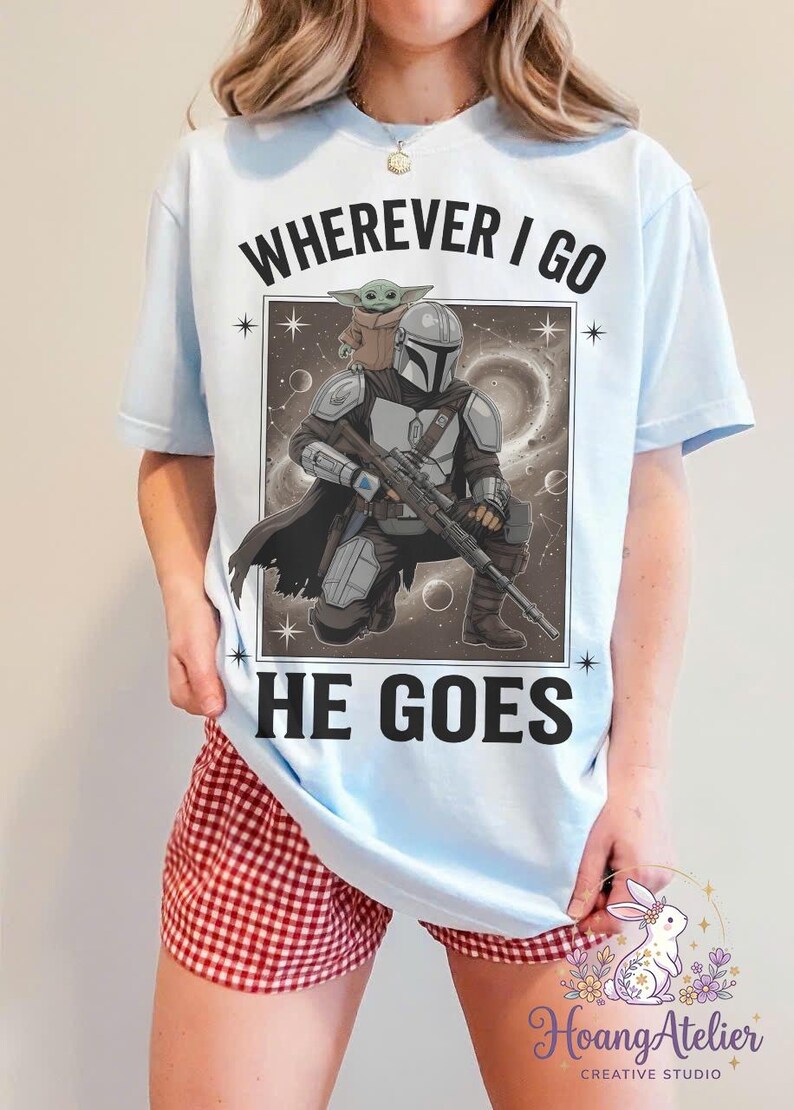 May include: Light blue t-shirt featuring a graphic of The Mandalorian and Grogu, with the text "WHEREVER I GO HE GOES". The design includes a detailed illustration of the characters in a space-themed background.