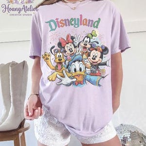 May include: A light purple t-shirt featuring the word "Disneyland" in colorful lettering, with a graphic of Disney characters including Mickey Mouse, Minnie Mouse, Donald Duck, and Goofy. The shirt is a relaxed fit.