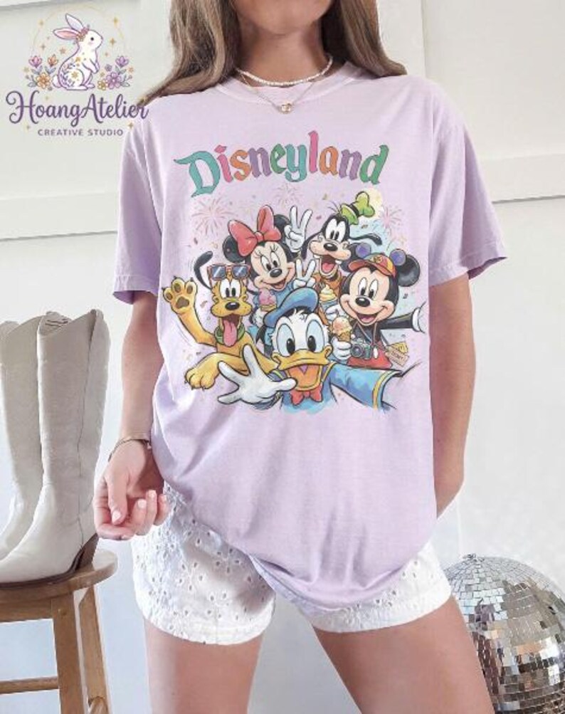May include: A light purple t-shirt featuring the word "Disneyland" in colorful lettering, with a graphic of Disney characters including Mickey Mouse, Minnie Mouse, Donald Duck, and Goofy. The shirt is a relaxed fit.