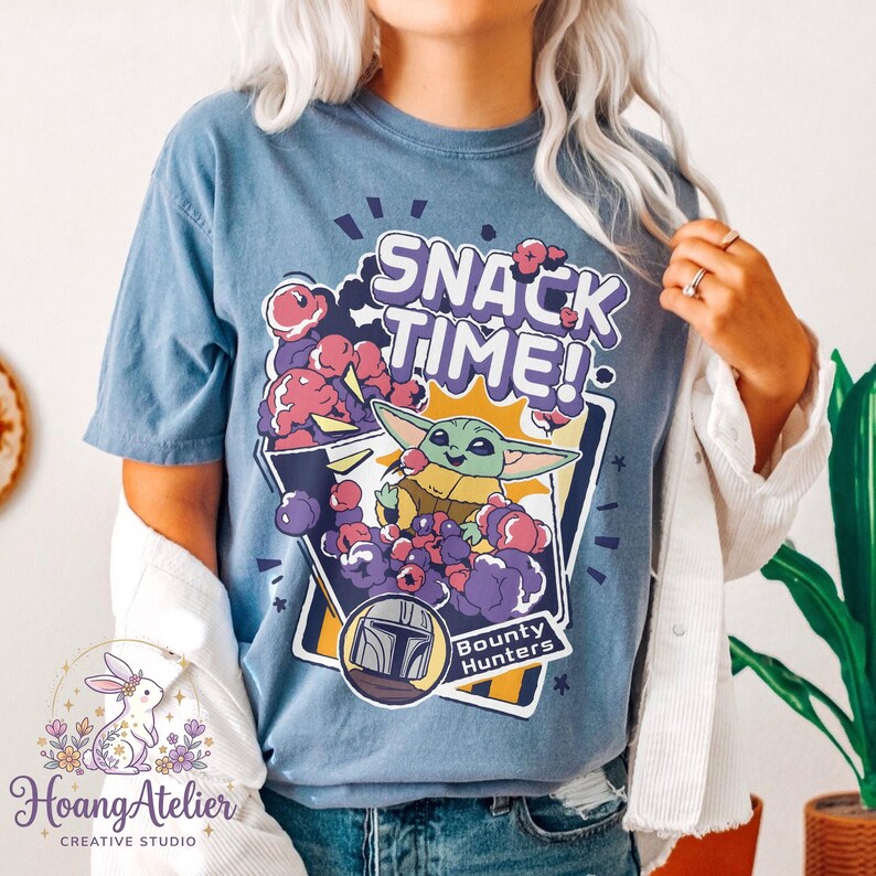 Puede incluir: Camiseta azul con un gr&aacute;fico de Grogu de The Mandalorian, rodeado de snacks morados y rosas. El texto "SNACK TIME!" est&aacute; encima de Grogu, con "Bounty Hunters" debajo. La camiseta tiene un corte holgado.