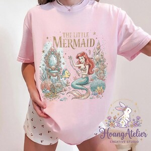 May include: Pink t-shirt featuring a colorful illustration of Ariel from The Little Mermaid. The graphic includes the text "THE LITTLE MERMAID" in gold lettering, along with underwater elements and a mermaid. The shirt is a light pink color.