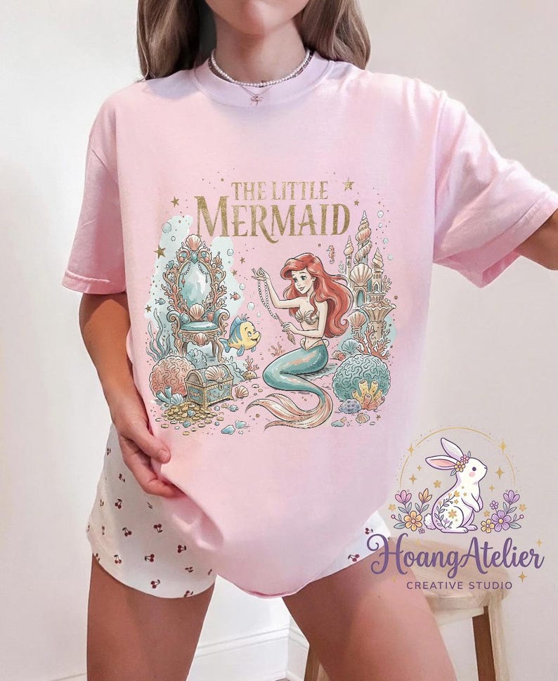 May include: Pink t-shirt featuring a colorful illustration of Ariel from The Little Mermaid. The graphic includes the text "THE LITTLE MERMAID" in gold lettering, along with underwater elements and a mermaid. The shirt is a light pink color.