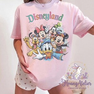 May include: A light pink t-shirt featuring the word "Disneyland" in a colorful, retro font. The shirt showcases a graphic of Disney characters including Mickey Mouse, Minnie Mouse, Goofy, Donald Duck, and Pluto. The shirt is a comfortable, casual style.