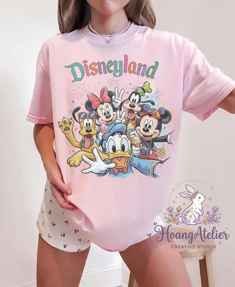 May include: A light pink t-shirt featuring the word "Disneyland" in a colorful, retro font. The shirt showcases a graphic of Disney characters including Mickey Mouse, Minnie Mouse, Goofy, Donald Duck, and Pluto. The shirt is a comfortable, casual style.