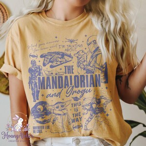 Puede incluir: Camiseta de color amarillo mostaza con ilustraciones y texto en azul de "The Mandalorian". El dise&ntilde;o incluye personajes, naves y frases como "This is the Way" y "Wherever I go, he goes."