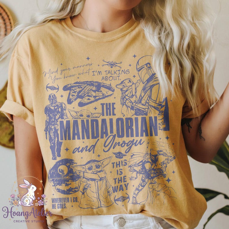 Puede incluir: Camiseta de color amarillo mostaza con ilustraciones y texto en azul de "The Mandalorian". El dise&ntilde;o incluye personajes, naves y frases como "This is the Way" y "Wherever I go, he goes."