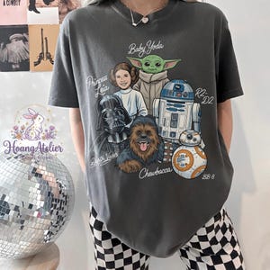 May include: Dark gray t-shirt featuring a graphic of Star Wars characters: Baby Yoda, Princess Leia, Darth Vader, Chewbacca, R2-D2, and BB-8. The text includes character names. The shirt is a comfortable, casual wear item.