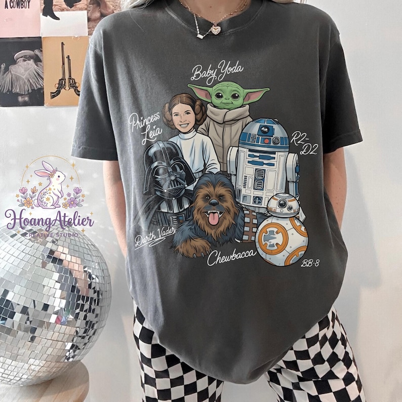 May include: Dark gray t-shirt featuring a graphic of Star Wars characters: Baby Yoda, Princess Leia, Darth Vader, Chewbacca, R2-D2, and BB-8. The text includes character names. The shirt is a comfortable, casual wear item.