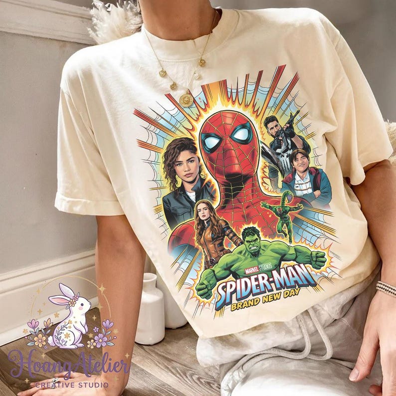 May include: Cream-colored t-shirt featuring a vibrant graphic of Spider-Man and other Marvel characters. The design includes the text "Spider-Man Brand New Day" in a dynamic, comic-book style. The shirt has a classic crew neck and short sleeves.