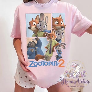 K&ouml;nnte beinhalten: Hellrosa T-Shirt mit einem Comic-Strip-Design mit Charakteren aus dem Animationsfilm "Zoomania". Das Design zeigt einen Fuchs, ein Kaninchen und andere Charaktere. Der Text "Zoomania 2" ist ebenfalls sichtbar.
