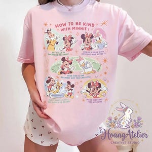 May include: Pale pink t-shirt with Minnie Mouse and friends, displaying the text "HOW TO BE KIND WITH MINNIE!". The design includes illustrations and phrases such as "Say hello to everyone you meet!" and "Make everyone feel welcome."