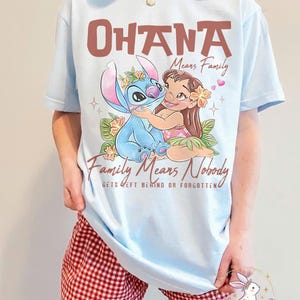 Op de afbeelding: Lichtblauw T-shirt met een afbeelding van Stitch en Lilo die elkaar omhelzen, met de tekst "OHANA Means Family" en "Family Means Nobody Gets Left Behind or Forgotten." Het ontwerp bevat bloemmotieven en harten.