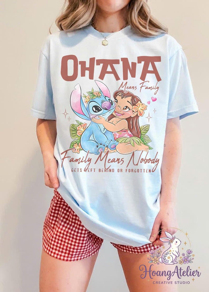 Op de afbeelding: Lichtblauw T-shirt met een afbeelding van Stitch en Lilo die elkaar omhelzen, met de tekst "OHANA Means Family" en "Family Means Nobody Gets Left Behind or Forgotten." Het ontwerp bevat bloemmotieven en harten.