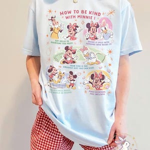 May include: Pale blue t-shirt featuring the text "HOW TO BE KIND WITH MINNIE!" and images of Minnie Mouse and friends. The shirt also has the text "SAY HELLO TO EVERYONE YOU MEET!" and "SHARE A SMILE WITH SOMEONE WHO NEEDS IT!"