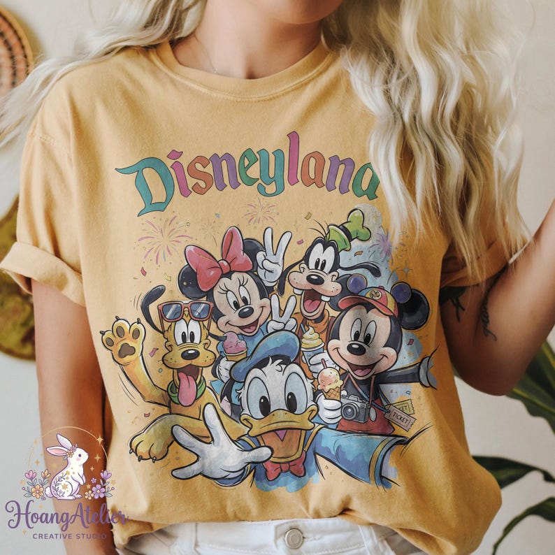 May include: Mustard yellow t-shirt featuring the word "Disneyland" in colorful text above a graphic of Mickey Mouse, Minnie Mouse, Donald Duck, Goofy, and Pluto. The characters are smiling and holding ice cream cones and a camera.