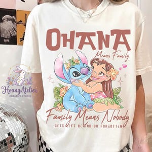 Op de afbeelding: Cr&egrave;mekleurig T-shirt met een afbeelding van Stitch en Lilo uit Lilo & Stitch, met de woorden "OHANA Means Family" en "Family Means Nobody Gets Left Behind or Forgotten." Het ontwerp bevat bloemaccenten en harten.