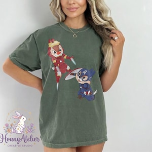 May include: Olive green t-shirt featuring cartoon characters dressed as superheroes. One character is dressed as Iron Man, and the other as Captain America. The shirt has the text "Hoang Atelier Creative Studio" at the bottom.
