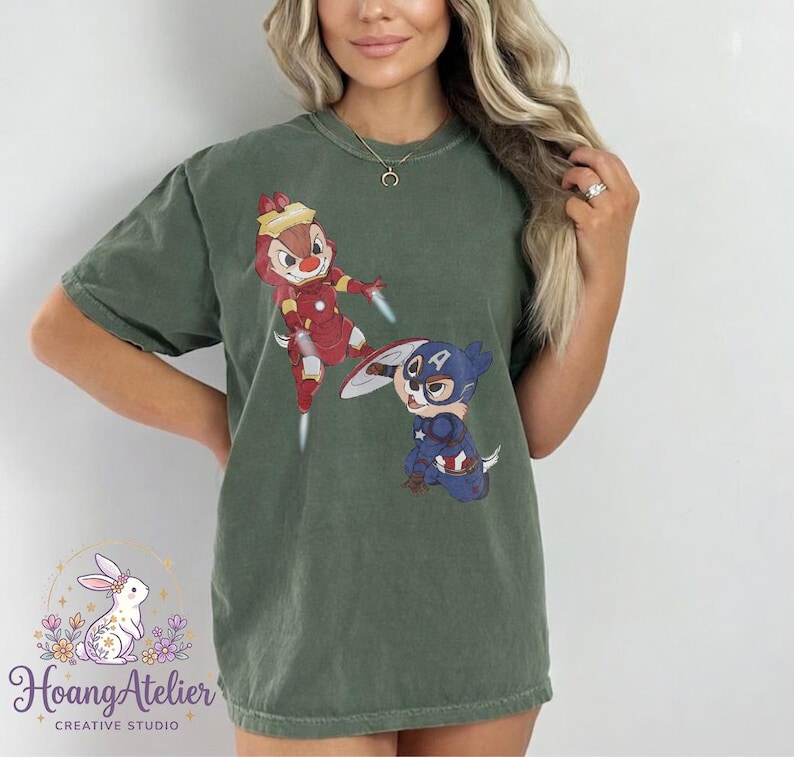May include: Olive green t-shirt featuring cartoon characters dressed as superheroes. One character is dressed as Iron Man, and the other as Captain America. The shirt has the text "Hoang Atelier Creative Studio" at the bottom.