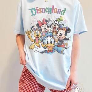 May include: Light blue t-shirt featuring a colorful Disneyland graphic with Mickey Mouse, Minnie Mouse, Donald Duck, Goofy, and Pluto. The word "Disneyland" is written in pastel colors above the characters. The shirt is paired with red and white checkered shorts.