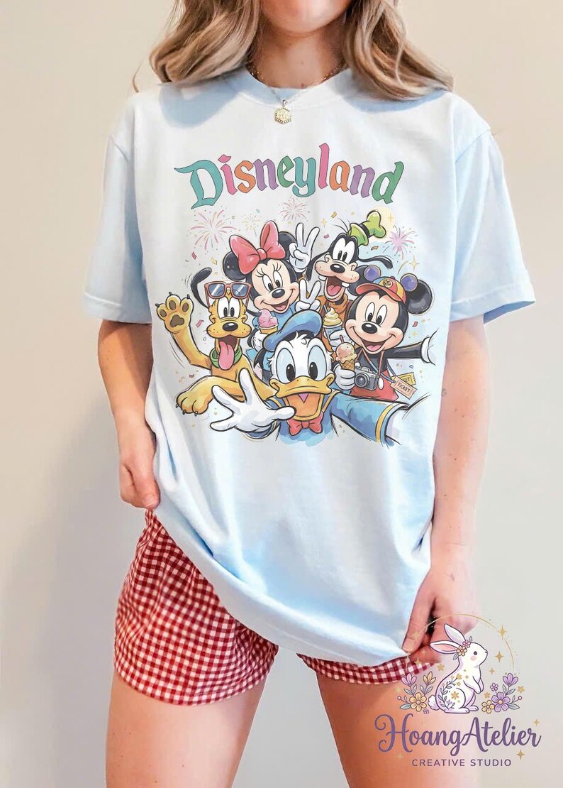 May include: Light blue t-shirt featuring a colorful Disneyland graphic with Mickey Mouse, Minnie Mouse, Donald Duck, Goofy, and Pluto. The word "Disneyland" is written in pastel colors above the characters. The shirt is paired with red and white checkered shorts.