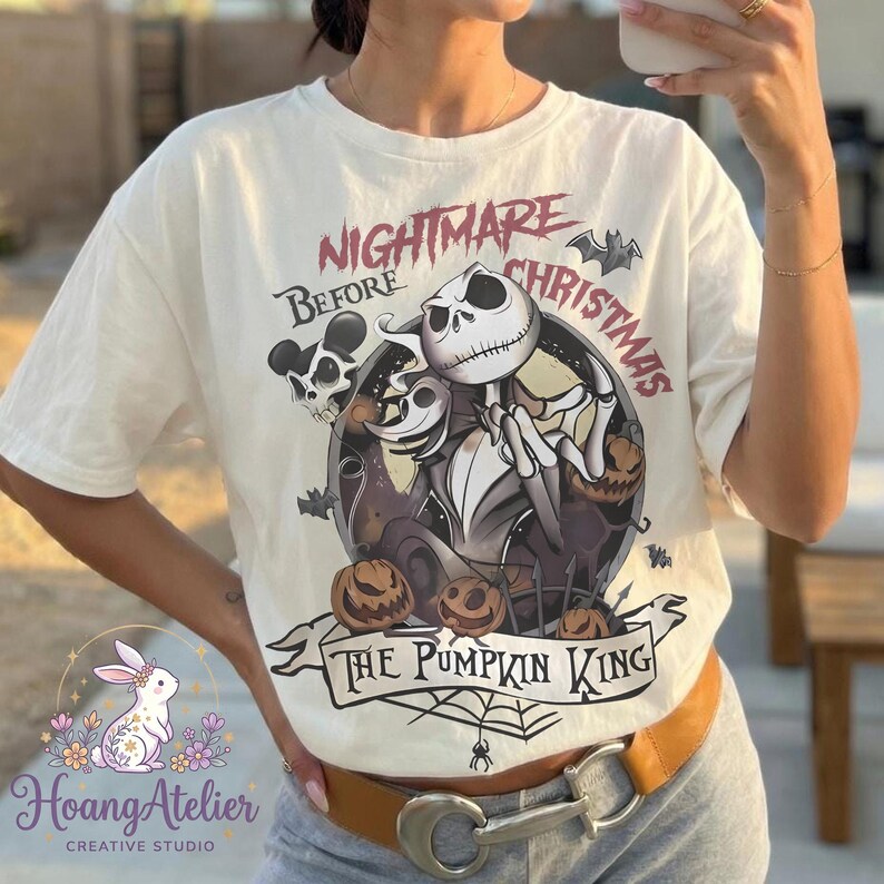 May include: Off-white t-shirt featuring a graphic from "The Nightmare Before Christmas." The design includes Jack Skellington, pumpkins, bats, and the text "Nightmare Before Christmas" and "The Pumpkin King." The shirt has short sleeves and a crew neck.