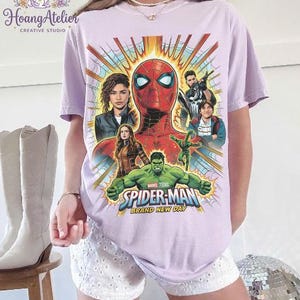 May include: A light purple t-shirt featuring a graphic of Spider-Man and other Marvel characters. The design includes the text "SPIDER-MAN BRAND NEW DAY" in blue and gold. The shirt has a crew neck and short sleeves.