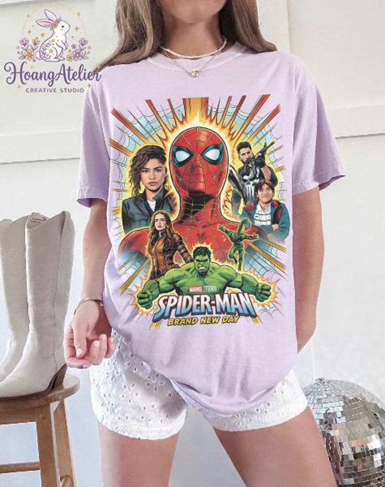 May include: A light purple t-shirt featuring a graphic of Spider-Man and other Marvel characters. The design includes the text "SPIDER-MAN BRAND NEW DAY" in blue and gold. The shirt has a crew neck and short sleeves.