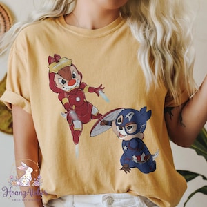 May include: Mustard yellow t-shirt featuring cartoon characters dressed as superheroes. One character is in a red Iron Man suit, and the other is in a Captain America suit. The shirt has rolled-up sleeves.