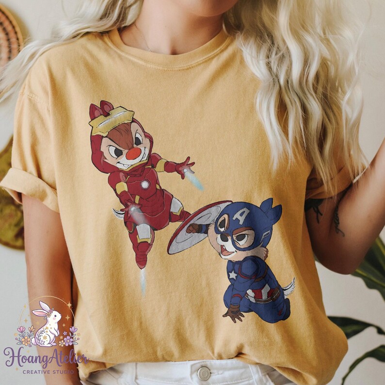 May include: Mustard yellow t-shirt featuring cartoon characters dressed as superheroes. One character is in a red Iron Man suit, and the other is in a Captain America suit. The shirt has rolled-up sleeves.