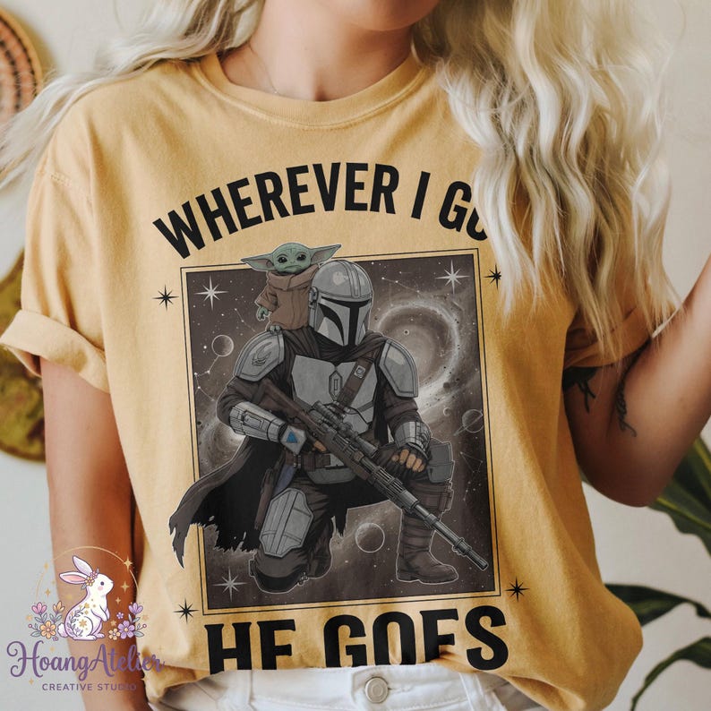May include: Mustard yellow t-shirt featuring a graphic of The Mandalorian and Grogu from the Star Wars series. The design includes the text "WHEREVER I GO HE GOES" in black, with a space-themed background. The shirt has rolled-up sleeves.